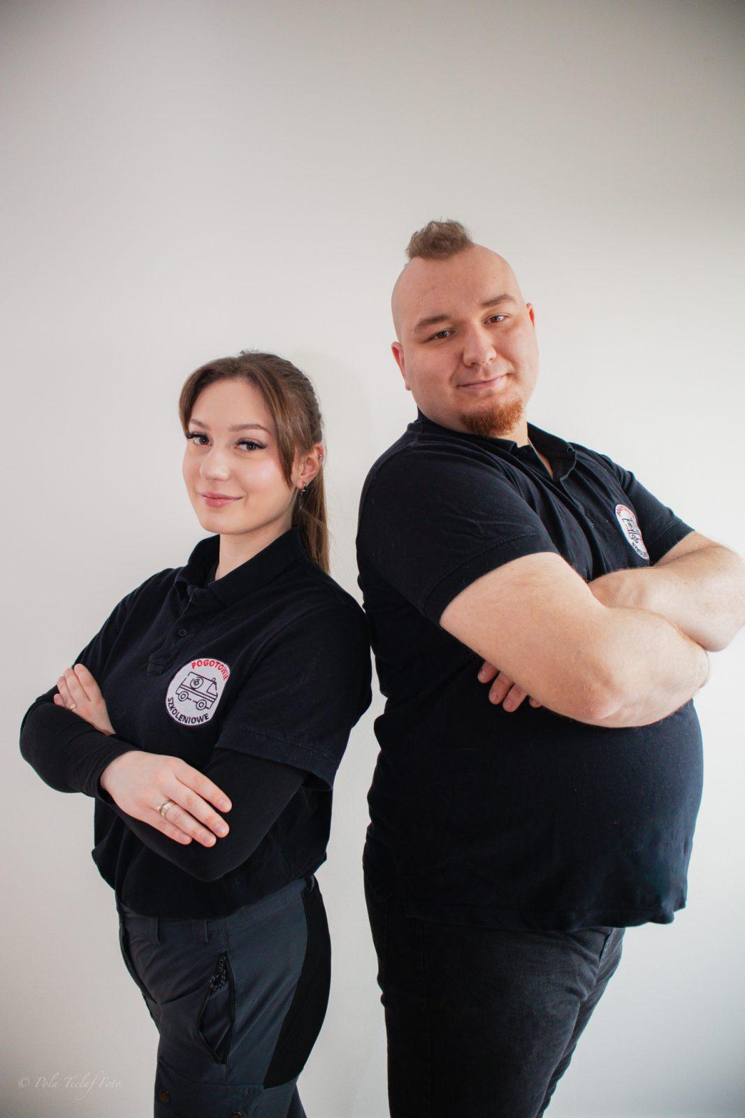 Dwóch instruktorów Pogotowia Szkoleniowego stojących tyłem do siebie z założonymi rękoma w czarnych koszulkach firmowych na jasnym tle